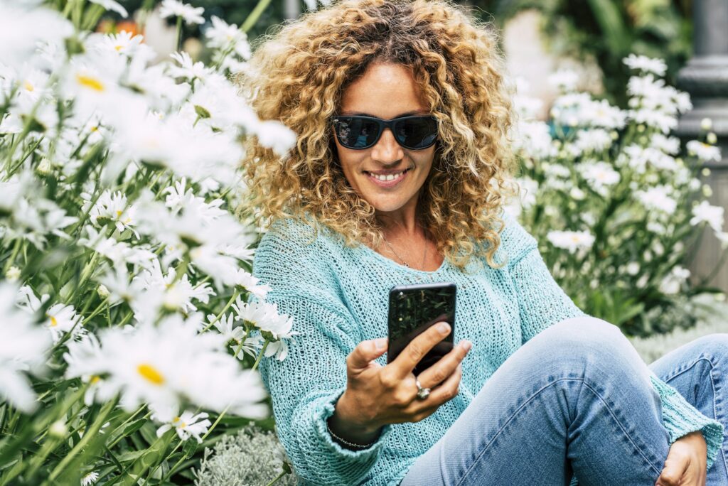 Cute caucasian woman using mobile phone outdoor sitting with daisies spring flowers around